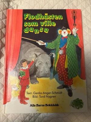 Flodhästen som ville dansa - En färgglad barnbok med hårda pärmar, skriven av Gerda Anger-Schmidt och illustrerad av Tord Nygren. Boken har en tecknad framsida med en clown, en flodhäst och ett barn, och är utgiven av Alla Barns Bokklubb. Perfekt för dig som gillar fantasifulla berättelser.