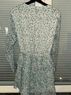 Blommig volangklänning med v-ringning - Söt kortklänning med grönt blommönster på vit botten, v-ringning och knappar framtill. Klänningen har långa ärmar och flera volanglager i kjolen för en flowy känsla. Perfekt för vår och sommar!