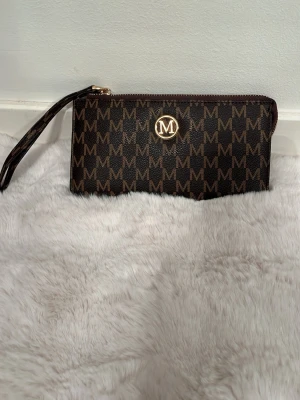 Brun plånbok/ mini väska - Snygg brun plånbok guldiga detaljer och M-logga över hela ytan. Plånboken har dragkedja och en praktisk handledsrem. Tillverkad i konstläder med klassiskt mönster. Perfekt för att hålla koll på kort och cash.