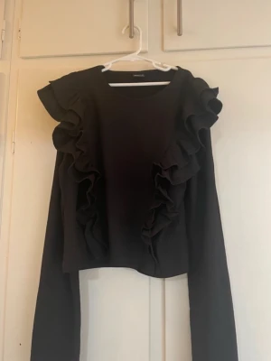 Svart topp med volangdetaljer - Snygg svart långärmad topp från Monki med volanger längs axlar och framsida. Rund halsringning och rak passform. Perfekt för dig som vill ha en trendig och stilren look med extra detaljer. Den passar som S dessutom och ej använd 