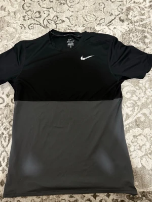 Nike Dri-Fit svart/grå träningströja - Snygg kortärmad träningströja från Nike i svart och grått. Tillverkad i Dri-Fit-material som andas och transporterar bort fukt. Perfekt för gymmet eller löprundan. Liten vit Nike-logga på bröstet och sportig design. Ganska liten i storleken så passar bättre för storlek S.