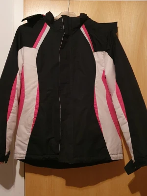 Vinterjacka - Snygg vinterjacka i svart med rosa och vita detaljer. Jackan har huva, dragkedja framtill och justerbara muddar vid ärmsluten. Fodrad för extra värme och har sportig design med färgblock längs sidorna. Perfekt för kalla dagar och utomhusaktiviteter. Storlek M för flicka. Har fläckar.