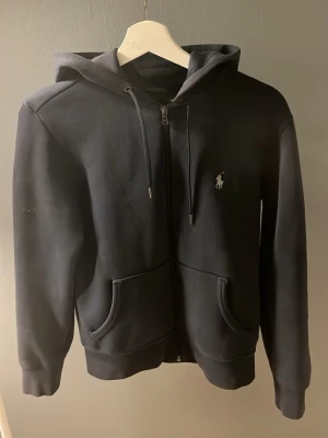 Svart hoodie från Ralph Lauren - Klassisk svart hoodie från Ralph Lauren med dragkedja, två fickor framtill och justerbar huva med snören. Ikoniska Polo-loggan broderad på bröstet. Tillverkad i mjukt bomullsmaterial som är skönt att ha på sig.