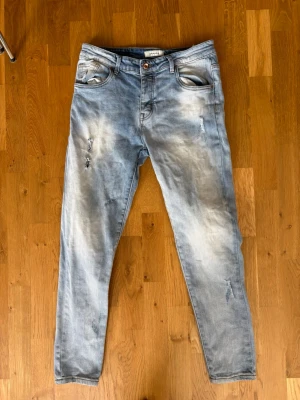 Ljusblå slitna jeans med skinny fit - Säljer ett par ljusblå jeans med skinny fit och slitna detaljer på benen. Jeansen har klassisk femficksdesign, knappgylf och är tillverkade i bomull med lite stretch för extra komfort. Perfekta för en avslappnad och trendig look.