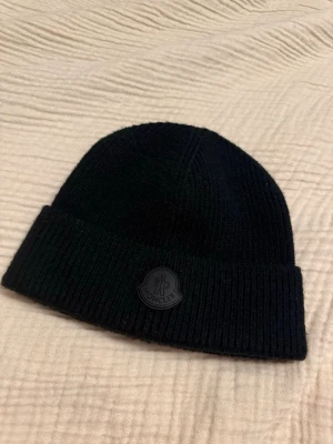 Moncler Mössa Svart Ull Beanie Logo One Size Äkta - Moncler mössa – svart 🖤  Säljer en stilren svart Moncler mössa i ribbad stickning med diskret gummilogo framtill. Klassisk modell som passar till allt och håller dig varm under höst och vinter.  Mössan är i mycket fint skick och sparsamt använd. QR-kod och etiketter finns kvar på insidan.  • Märke: Moncler • Färg: Svart • Storlek: One size • Skick: Väldigt bra  Nypris ca 2500–3000 kr.  Pris: 1200 kr (kan diskuteras vid snabb affär).  Skickas eller möts upp om det passar.
