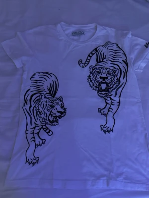 Vit Kenzo t-shirt - Vit t-shirt från Kenzo med två stora svarta tigermotiv framtill. Klassisk rund halsringning och korta ärmar. Perfekt för dig som vill sticka ut med en ikonisk design från ett välkänt märke.