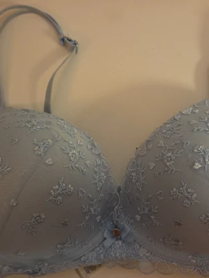 Ljusblå spets-bh med broderi - Söt ljusblå push-up bh med vadderade kupor och justerbara axelband. Utsidan är dekorerad med blommigt spetsbroderi och en liten rosett framtill. Bh:n har bygel och fina spetsdetaljer längs kanterna för en romantisk look. Insidan är sliten med utsidan ser fin ut. Priset går att diskutera!