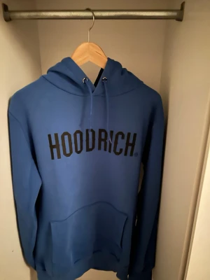 Blå HOODRICH hoodie med huva - Säljer en blå hoodie från HOODRICH med stor svart logga på bröstet. Hoodien har huva med dragsko och en stor magficka framtill. Tillverkad i mjukt material, perfekt för en chill och avslappnad stil.