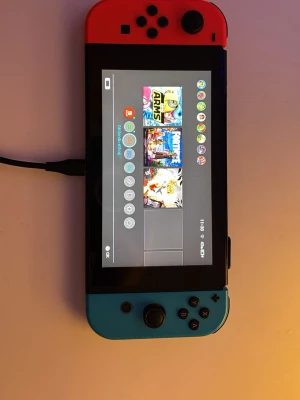 Nintendo Switch med neonröda och blå Joy-Con - Nintendo Switch spelkonsol med en svart kropp och avtagbara Joy-Con-kontroller i neonrött och neonblått. Konsolen har en rektangulär form och stor skärm i mitten. Perfekt för både handhållet och dockat spelande. Materialet är plast.