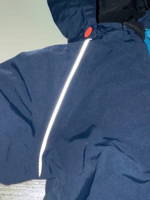 Mörkblå vindjacka från Polarn O. Pyret - Snygg mörkblå vindjacka med huva från Polarn O. Pyret. Jackan har turkos dragkedja, reflexdetaljer, röda knappar och en liten orange detalj vid bröstet. Perfekt för blåsiga dagar och enkel att matcha med andra plagg. Polyamid o polyester