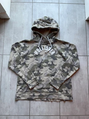 Camo hoodie från Hollister - Snygg hoodie från Hollister i camomönster med texten 'Hollister Los Angeles' på bröstet. Utrustad med huva och vita dragsnören samt en stor magficka. Perfekt för en avslappnad streetstyle-look.