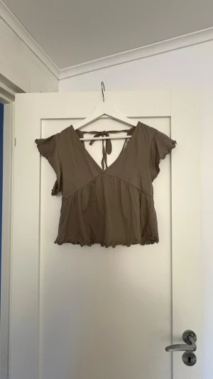 Brun croppad topp med knyt i ryggen - Säljer en brun croppad topp med v-ringning och korta ärmar. Toppen har ett sött knyt i ryggen och volangkant nedtill. Perfekt till sommaren och enkel att styla med jeans eller kjol.