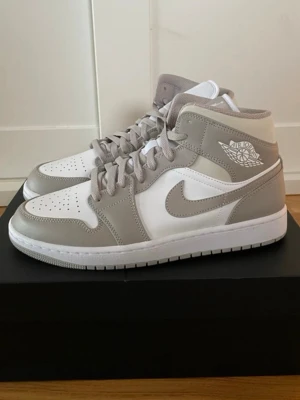 Air Jordan 1 Mid Vit/Grå - Snygga Air Jordan 1 Mid sneakers i vitt och ljusgrått läder. Klassisk siluett med perforerad tå, vita snören och ikoniska Air Jordan-loggan på sidan. Perfekta för dig som vill ha en clean och stilren look med streetkänsla.
