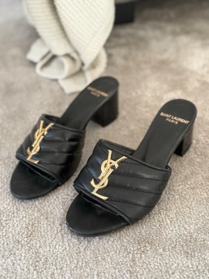 YSL Svarta klackar  - Säljer ett par svarta klackar med breda remmar i quiltat läder. Skorna har öppen tå och en chunky klack som ger en cool vibe. Perfekta för dig som vill ha något lyxigt och trendigt. Liten skada framtil. 