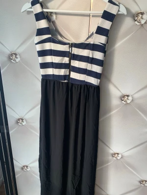 Randig klänning med svart kjol - Snygg ärmlös klänning med marinblå och vita horisontella ränder upptill och enfärgad svart kjoldel. Klänningen har breda axelband och dragkedja bak. Överdelen är i ribbat material och kjolen i ett mjukare tyg. Perfekt för en trendig och avslappnad look.