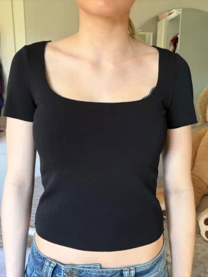 Svart croppad topp med fyrkantig ringning - Snygg svart croppad topp med fyrkantig ringning och korta ärmar. Perfekt till jeans eller kjol för en trendig look. Tunn och stretchig kvalitet som sitter tight och framhäver figuren.