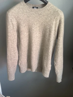 Grå stickad tröja Uniqlo - Klassisk beige stickad tröja från Uniqlo med rund halsringning och långa ärmar. Tröjan har en enkel och stilren design, perfekt för lager på lager. Tillverkad i mjukt material som känns skönt mot huden.