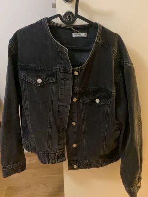 Svart jeansjacka utan krage - Cool svart jeansjacka med rund halsringning och silverfärgade knappar. Klassisk modell med två bröstfickor med lock och knapp. Långärmad och i ett mjukt denimtyg som ger en avslappnad vibe.