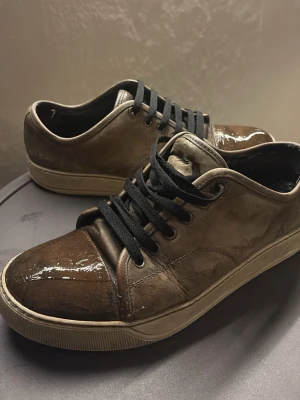 Bruna sneakers från Lanvin i läder - Snygga bruna sneakers från Lanvin i glansigt läder med svarta skosnören och beige sula. Skorna har en låg profil och en stilren design som passar till många outfits. Perfekta för dig som gillar exklusiva och trendiga sneakers.