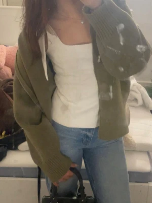 Olivgrön stickad hoodie - Mysig olivgrön stickad hoodie med dragkedja och huva med vita snören. Tröjan har en avslappnad passform och ribbade muddar vid ärmslut och nederkant. Perfekt för en chill och avslappnad stil.