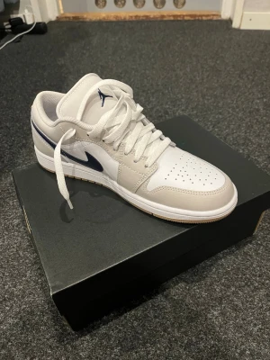 Air Jordan 1 Low - Modell: Air Jordan 1 low Färg: vit, grå, navy blå, brun/beige sula  Helt nya och oanvända. Har endast satt i snöre i ena skon för visualitet. Original kartong medföljer. Nypris: 1499kr Denna färgkombination går ej köpa direkt ifrån Nike längre.