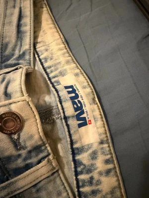 Mavi Jeans - Säljer ett par ljusblå acid wash jeans från Mavi med klassisk femficksmodell och knappgylf. Jeansen har en unik tvätt med blåa detaljer och är tillverkade i bomull för en skön och avslappnad look. Perfekta för dig som gillar streetwear och vill sticka ut.