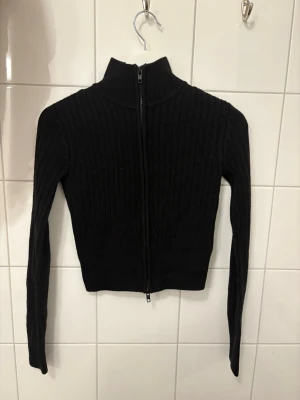 Svart ribbstickad ziptröja H&M - Svart ribbstickad tröja från H&M med dragkedja framtill och hög krage. Tröjan är figurnära och har långa ärmar, perfekt för lager-på-lager-stil. Enkel och stilren design som funkar till många outfits.