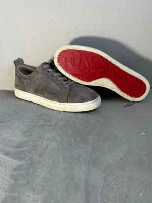 Gråa sneakers med röd sula - Snygga gråa sneakers från Christian Louboutin i mocka med klassisk röd sula och vita kanter. Skorna har lågt skaft, rund tå och matchande grå skosnören. Perfekta för dig som vill sticka ut med en ikonisk detalj under fotenv.