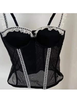 Svart spetskorsett med vita detaljer - Snygg svart korsett i transparent mesh med vita spetsdetaljer längs kupor och axelband. Justerbara axelband och dekorativa vertikala spetsband framtill. Perfekt för att skapa en cool och feminin look.
