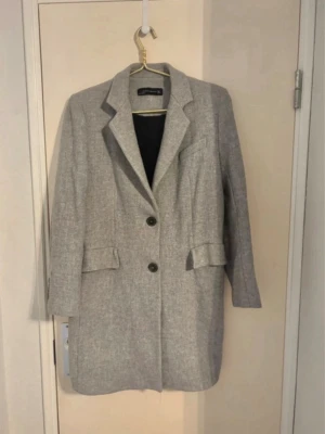 Ljusgrå klassisk ullrock - Snygg ljusgrå rock från Zara Basic med två knappar framtill, klassisk krage och två fickor med lock. Rak passform och helfodrad insida. Perfekt för dig som gillar stilrena och tidlösa plagg. Materialet känns som ullblandning.. Ny utan prislapp, inga skador eller fläckar 