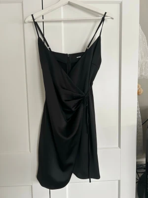 Svart omlottklänning med smala axelband - Säljer en svart kort omlottklänning från Bikbok med smala axelband och knyt i sidan. Klänningen har en draperad effekt framtill och en elegant v-ringning. Perfekt för dig som gillar stilrena och trendiga plagg.