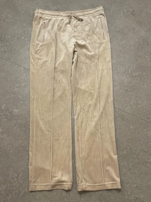 Beige velourbyxor Juicy Couture - Säljer ett par beige mjukisbyxor från Juicy Couture i mjuk velour. Byxorna har raka ben, dragsko i midjan och fickor med dragkedja fram. På baksidan finns Juicy-loggan i strass. Perfekta för en chill och trendig look. Kan mötas upp i Stockholm.