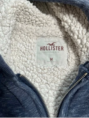 Hollsiter hoodie med dragkedja  - Mysig blåmelerad hoodie från Hollister med dragkedja och vit teddyfoder på insidan. Klassisk huva med dragsko och två fickor framtill. Perfekt för dig som gillar en avslappnad och varm stil. Broderad Hollister-logga på bröstet.