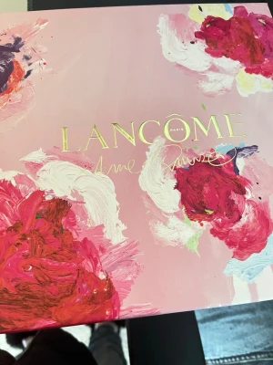 Lancôme La Vie Est Belle presentbox - Säljer en exklusiv presentbox från Lancôme med parfymen La Vie Est Belle, body lotion, en miniparfym och mascara. Boxen har ett konstnärligt blommigt rosa motiv och guldfärgad text. Perfekt för dig som älskar lyxiga skönhetsprodukter och vill ha allt i ett snyggt paket.