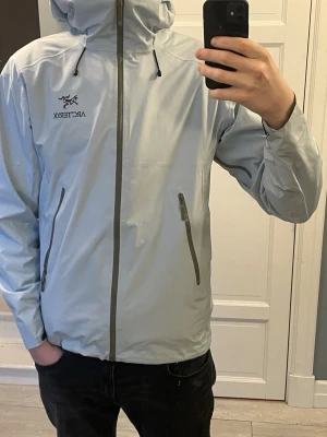 Ljusgrå vindjacka från Arc'teryx - Snygg ljusgrå vindjacka från Arc'teryx med huva. Jackan har två snedställda fickor med dragkedja och justerbara snören vid huvan. Tillverkad i ett tunt, vattenavvisande material som passar perfekt för blåsiga dagar. Perfekt för våren. Passar dig som är 190+ XL men sitter mer som L