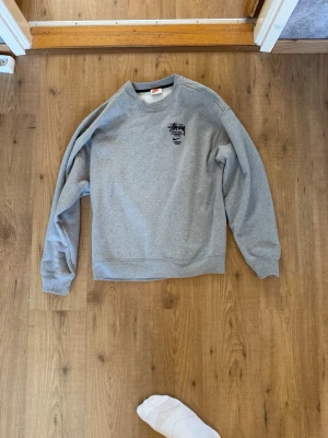 Nike x Stussy Sweatshirt - En stilren grå långärmad Nike X Stussy tröja med svart logga tryckt på bröstet. Tröjan har rund halsringning och långa ärmar med muddar. Perfekt för en avslappnad och trendig look. Storlek M. Pris kan diskuteras vid snabb affär.