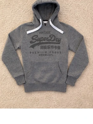 Grå Superdry hoodie med tryck - Snygg grå hoodie från Superdry med stort vintage-tryck på bröstet och japanska tecken. Klassisk känguruficka framtill, vita dragsnören och ribbade muddar. Perfekt för en avslappnad streetstyle-look.