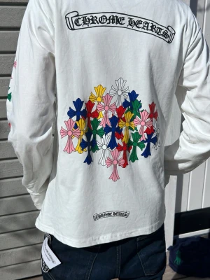 Vit långärmad Chrome Hearts t-shirt - Vit långärmad t-shirt från Chrome Hearts med färgglada blommotiv på ryggen och ärmarna. Bröstficka med logga framtill och coola detaljer i flera färger. Tillverkad i mjuk bomull och perfekt för dig som gillar streetwear och unika prints.