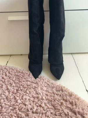 Svarta knähöga mockastövlar från lululia - Säljer ett par svarta knähöga stövlar i mocka med spetsig tå och låg klack. Stövlarna har en elegant siluett och en liten gulddetalj upptill på skaftet. Perfekta för dig som vill ha en stilren och trendig look.
