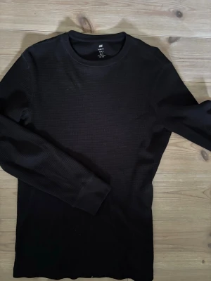 Svart långärmad tröja H&M Slim Fit - Svart långärmad tröja från H&M i slim fit-modell. Tröjan har rund halsringning och är tillverkad i ett strukturerat, ribbat material som ger en cool och enkel look. Perfekt att styla med jeans eller under en jacka.