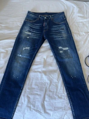 Mörkblå distressed jeans Dsquared2 - Snygga mörkblå jeans från Dsquared2 med slitningar, färgstänk och lätt tvättad look. Klassisk femficksmodell med raka ben och låg midja. Perfekta för en trendig streetstyle. Materialet är bomull med stretch för extra komfort.