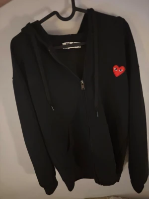 Svart hoodie Comme des Garçons Play - Svart hoodie med dragkedja från Comme des Garçons Play. Klassisk design med huva och snörning, samt det ikoniska röda hjärtat med ögon broderat på bröstet. Perfekt för en avslappnad och trendig stil. Sitter liten rekommendation på folk som är ungefär 170-175