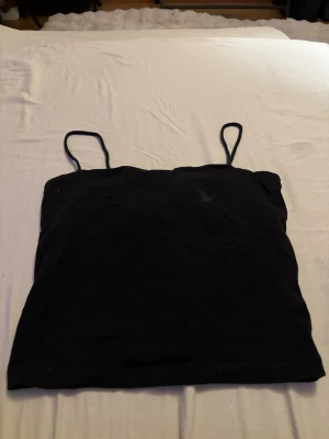 Svart linne med smala axelband - Säljer ett enkelt svart linne med smala axelband. Perfekt att ha under en skjorta eller bara som det är till jeans eller kjol. Linnet är figurnära och har en klassisk rak modell. Materialet känns mjukt och stretchigt.