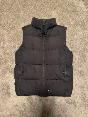 Svart dunväst från Champion - Snygg svart dunväst från Champion med hög krage och dragkedja framtill. Västen har två fickor med knappstängning på sidorna och quiltad design som ger en sportig look. Perfekt att slänga över en hoodie för extra värme.