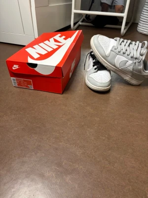 Nike Dunk Low vita sneakers - Nike Dunk Low sneakers i vitt och ljusgrått läder med klassisk swoosh på sidorna. Ena skon har svarta skosnören och den andra vita, vilket ger en unik touch. Skorna har perforerad tåbox och låg silhuett. Perfekta för dig som gillar streetwear och stilren design.