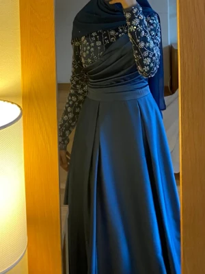 Blå långklänning med spetsdetaljer - Elegant blå långklänning med transparenta långärmade spetsärmar dekorerade med blommiga broderier och pärlor. Klänningen har draperad överdel och slät kjol i satinliknande material. Perfekt för speciella tillfällen. Använt en gång men kan vara smutsig på botten.