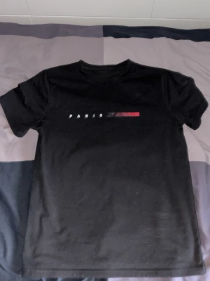 Svart t-shirt med Paris-tryck - Snygg svart t-shirt med texten 'PARIS' och en färgad linje i vitt, grått och rött på bröstet. Klassisk rak passform och rund halsringning. Perfekt till jeans eller shorts för en clean look.