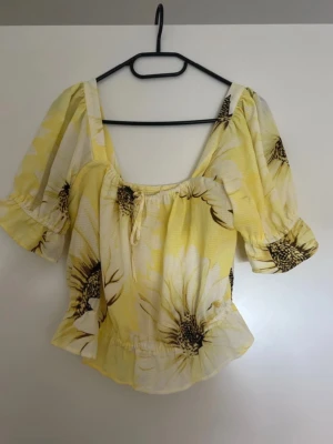 Gul blommig blus med volang - Superfin gul blus med stora vita blommor och bruna detaljer. Blusen har korta puffärmar med volangkant, fyrkantig urringning och smockad rygg. Lätt och luftig, perfekt för varma dagar. Söt knytning framtill och volang nertill ger en romantisk vibe.