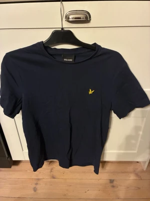 Mörkblå t-shirt från Lyle & Scott - Snygg mörkblå t-shirt från Lyle & Scott med klassisk passform och gul logga broderad på bröstet. T-shirten har rund halsringning och är tillverkad i mjuk bomull, perfekt för en stilren och enkel look. Använd ett fåtal gånger och har storleken M
