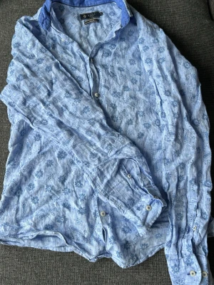 Blommig blå linneskjorta från Riley - Snygg ljusblå skjorta från Riley i linne med blommigt mönster och klassisk krage. Slim fit-modell med långa ärmar och vita knappar. Perfekt för dig som gillar en avslappnad men ändå stilren look.